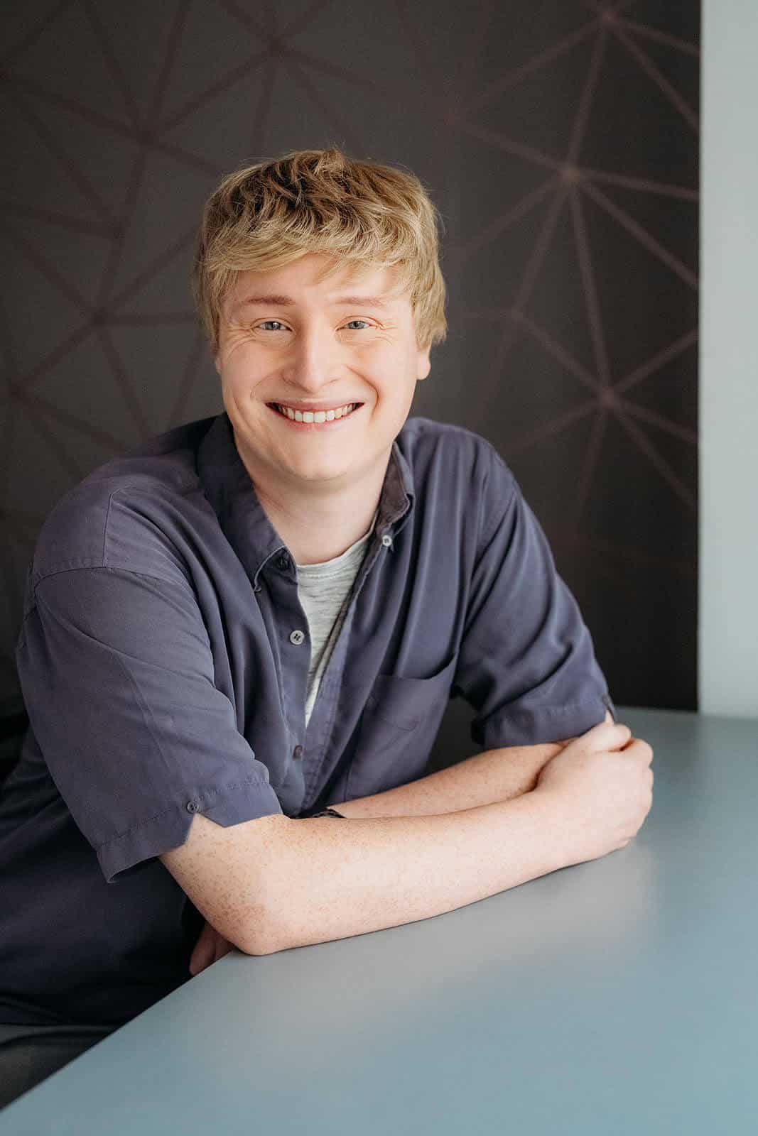 A person with short blond hair and a friendly smile sits at a table. They are wearing a dark gray shirt over a lighter gray top. The background features a geometric pattern on a dark wall.