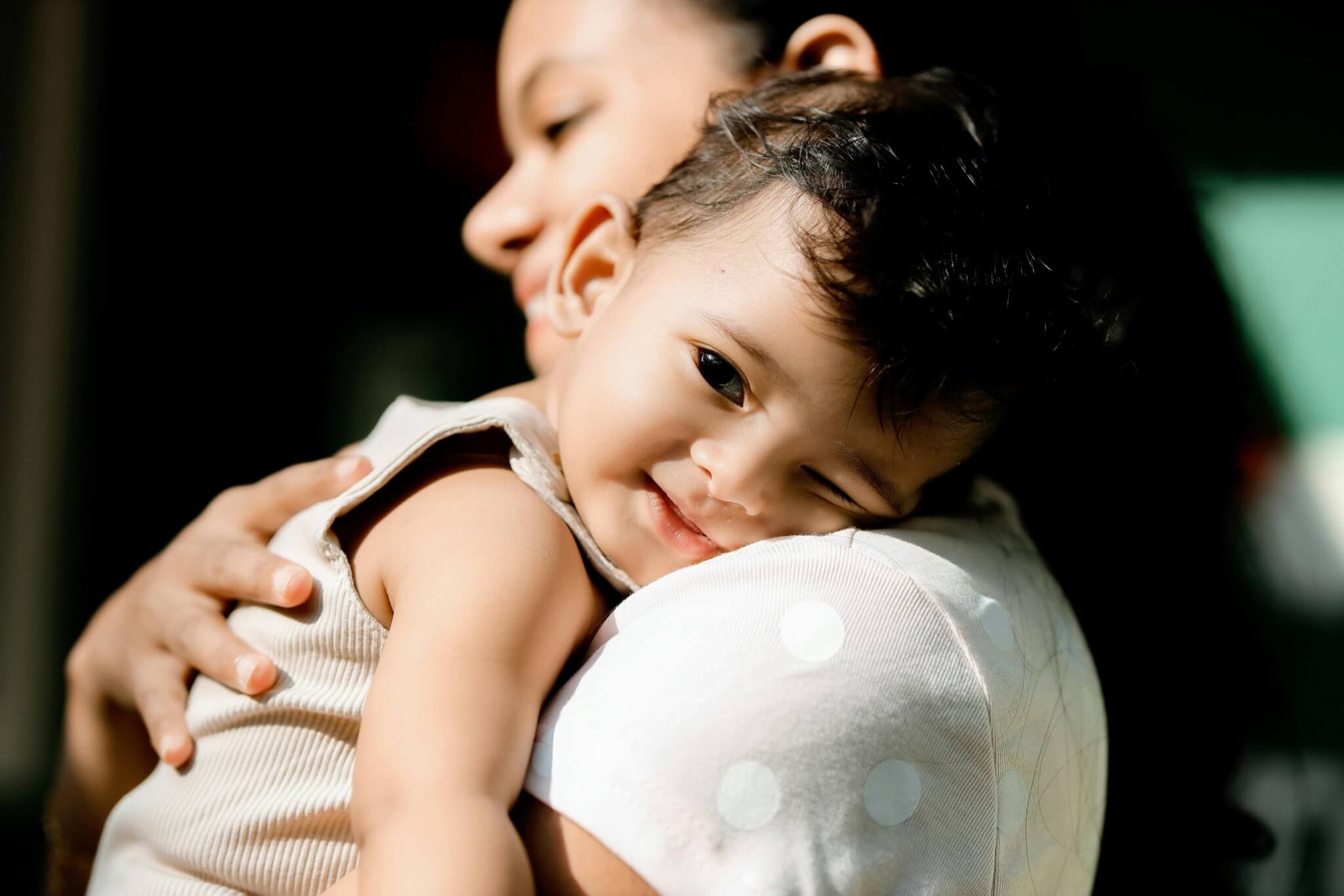 A smiling baby winks while being held lovingly by an adult, a heartwarming scene that brings light to overcome postnatal depression. The baby is wearing a light-colored outfit, and the softly blurred background emphasizes this warm, joyful moment.