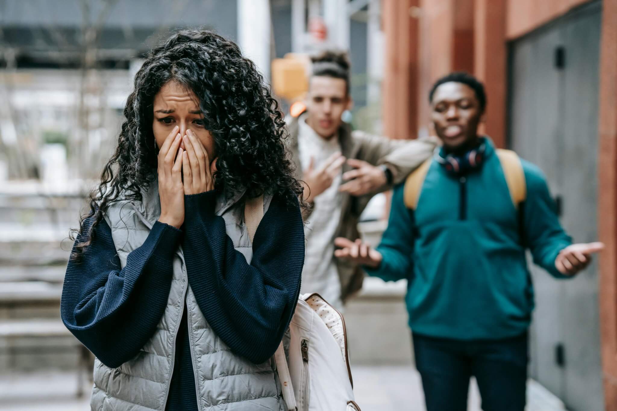 A young woman with curly hair looks distressed, holding her hands to her face, suggesting the weight of a painful secret. Behind her, two young men gesture animatedly in an urban setting, unaware of her struggle with the trauma of sexual abuse.