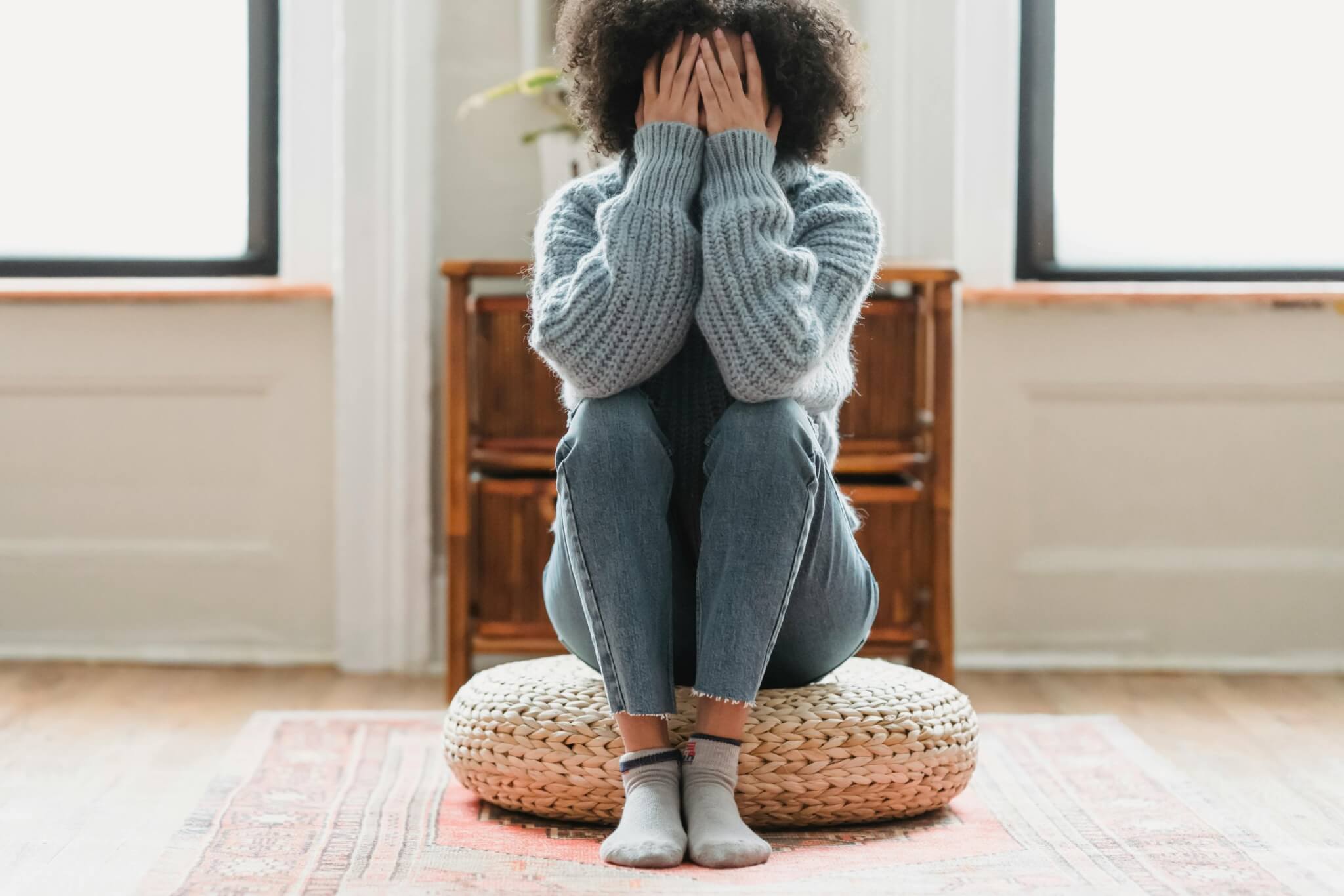 A person sits on a round pouf with their knees drawn up, wearing a cozy gray sweater and jeans. Their face is covered with their hands, perhaps reflecting the weight of postnatal depression. The room has warm lighting, a decorative rug, and large windows in the background.
