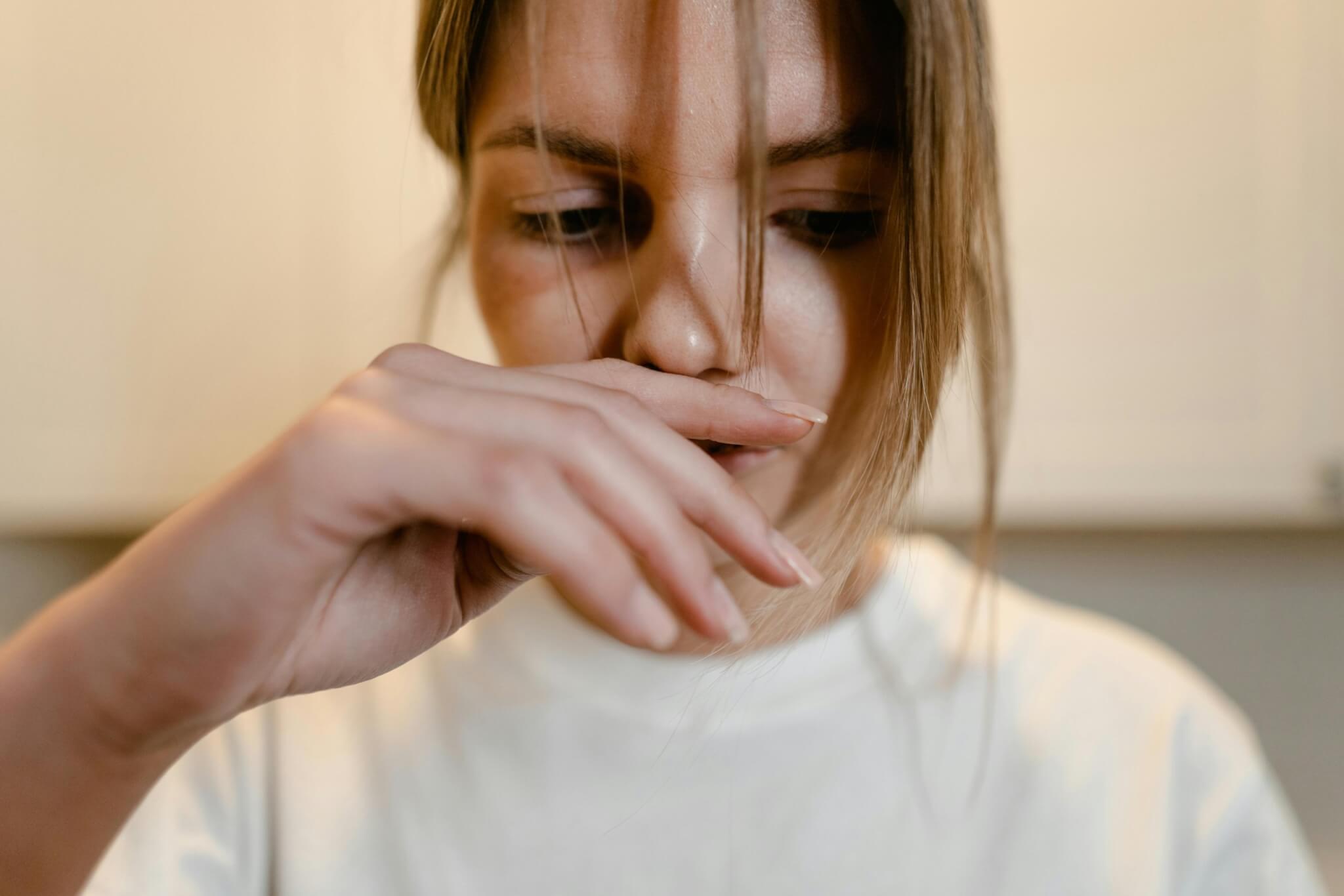 A woman with long hair partly covering her face looks down, her hand drawn close to her nose and mouth. Clad in a white top, she exudes a sense of contemplation amidst the softly blurred background, subtly hinting at the silent echoes of sexual abuse.