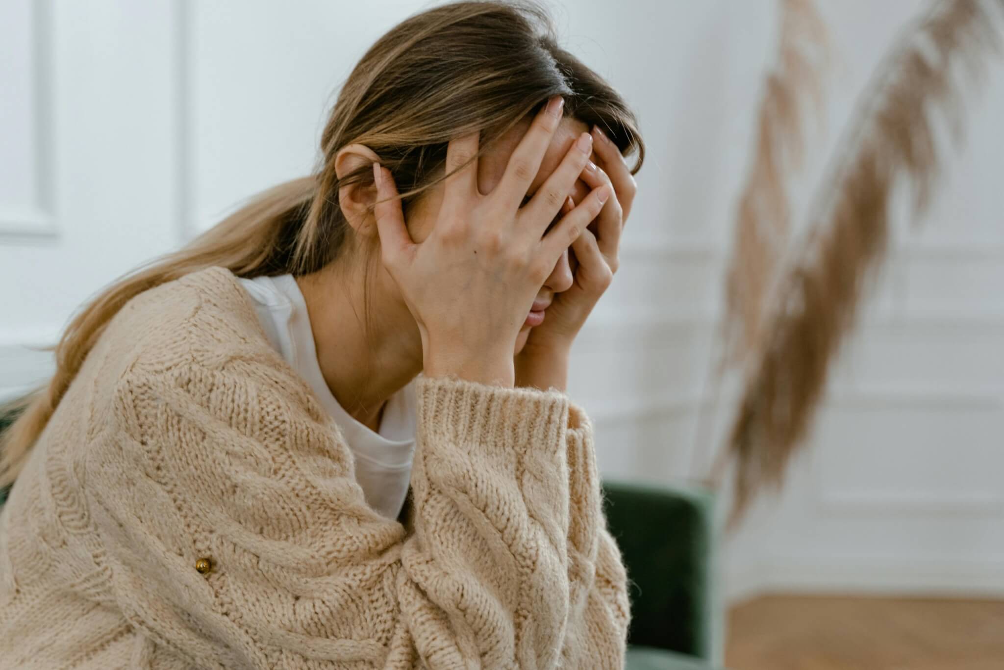 A woman sits in a room, her hands covering her face, embodying the weight of frustration or stress. Clad in a beige knitted sweater, she battles the silent aftermath of sexual abuse, with dried plants hazily silhouetted in the background.