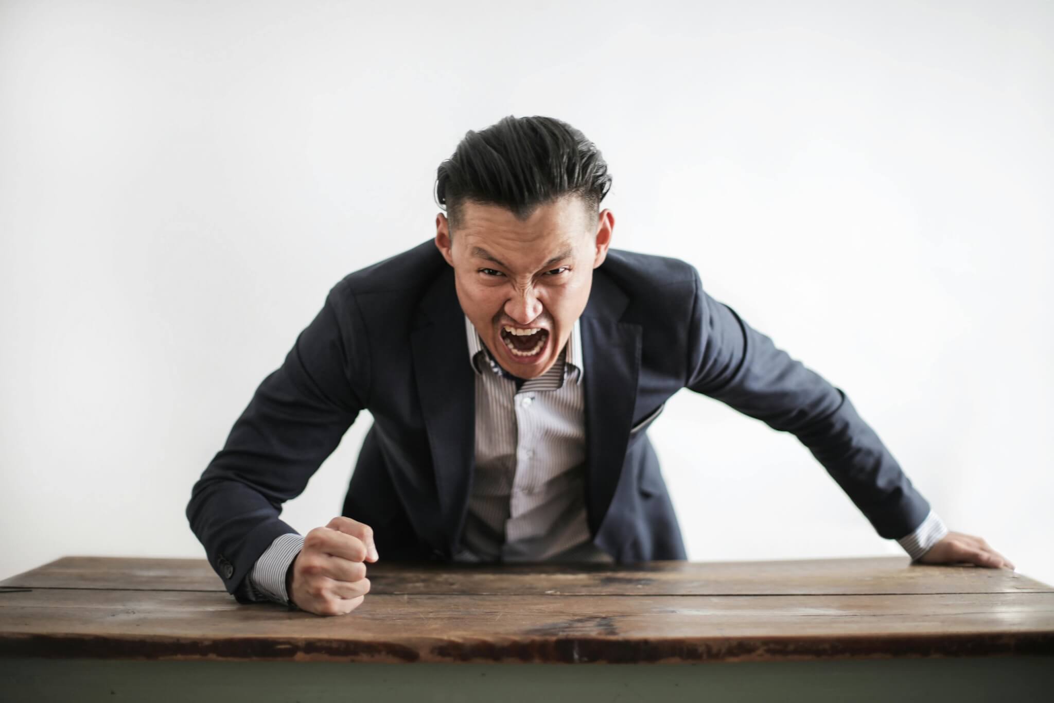 A man in a suit leans over a table, expressing anger with an open mouth and clenched fist, embodying the intensity often explored in therapy sessions, against a plain background.