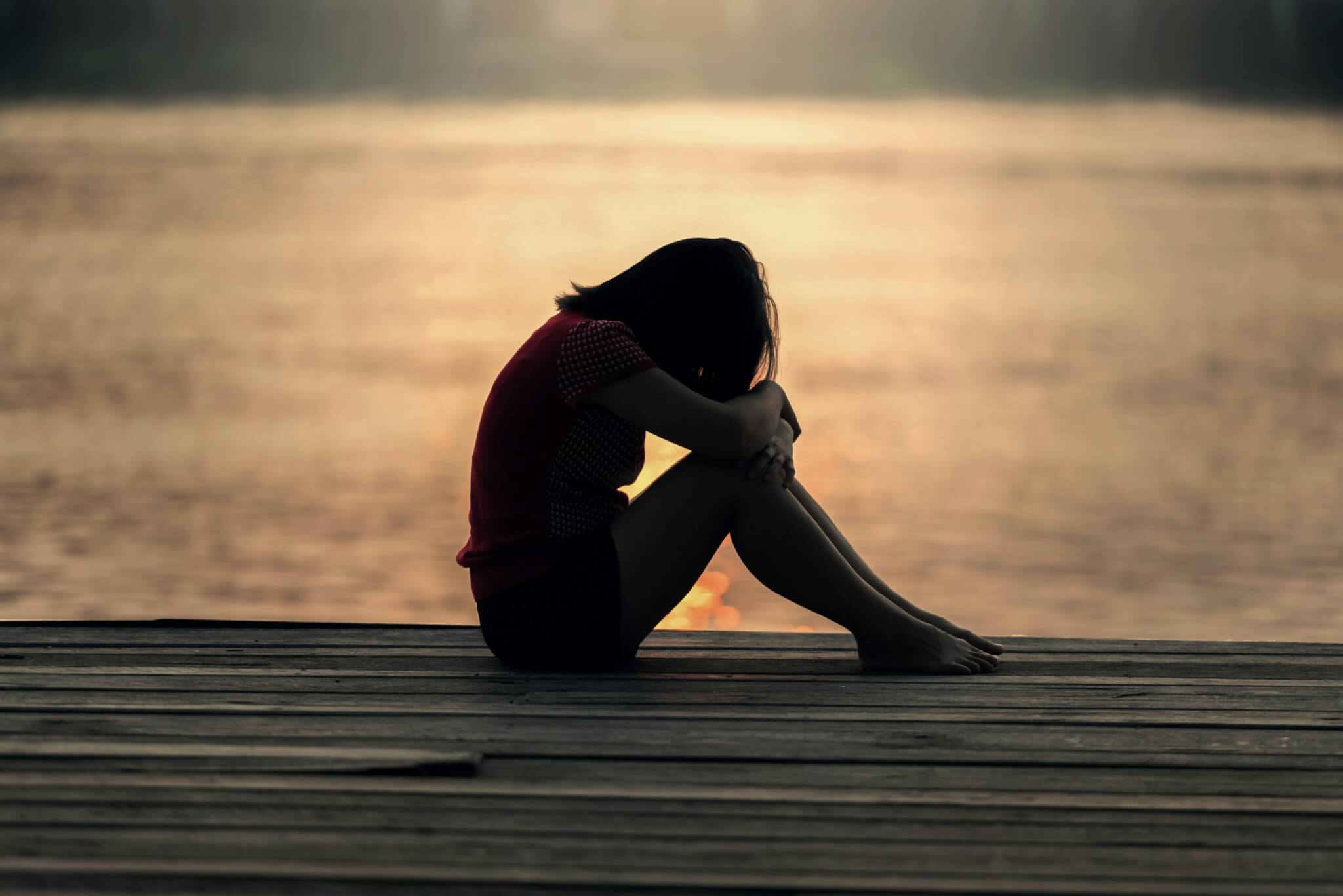Silhouette of a person sitting with knees drawn up and head resting on arms, on a wooden dock by a body of water during sunset. The warm glow reflects on the waters surface, offering solace in a serene atmosphere—a quiet moment away from the struggles of postnatal depression.