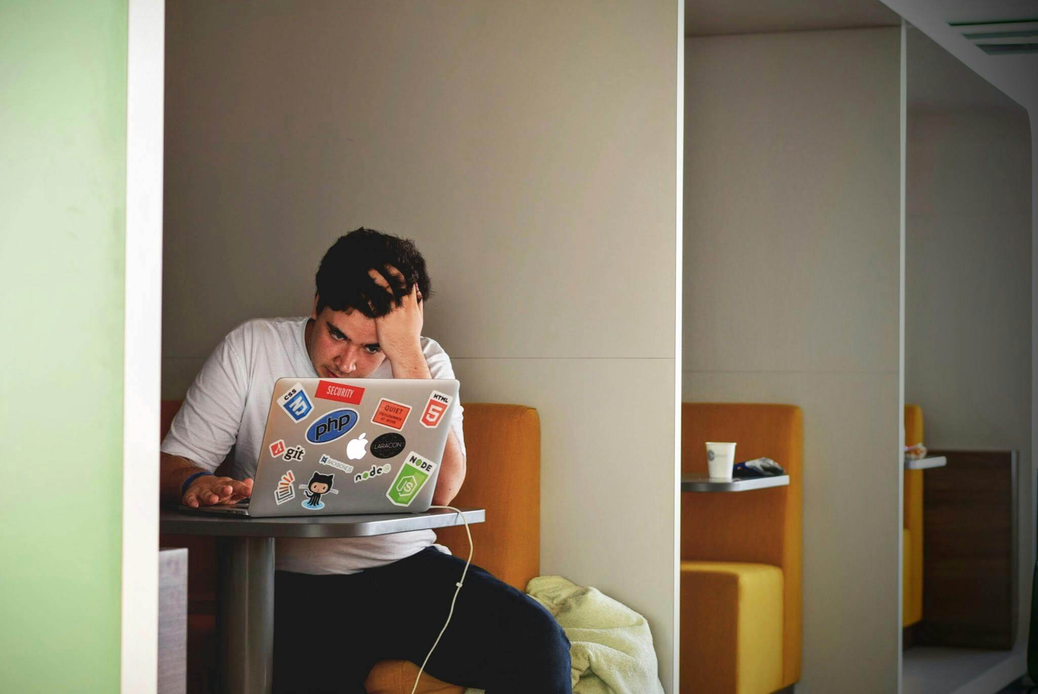 In a Surrey workspace, a person in a white shirt sits at a booth, deeply focused on their laptop adorned with tech stickers. One hand rests on their forehead, suggesting they might be pondering over conditions related to therapy. A cup sits nearby, adding to the casual setting.