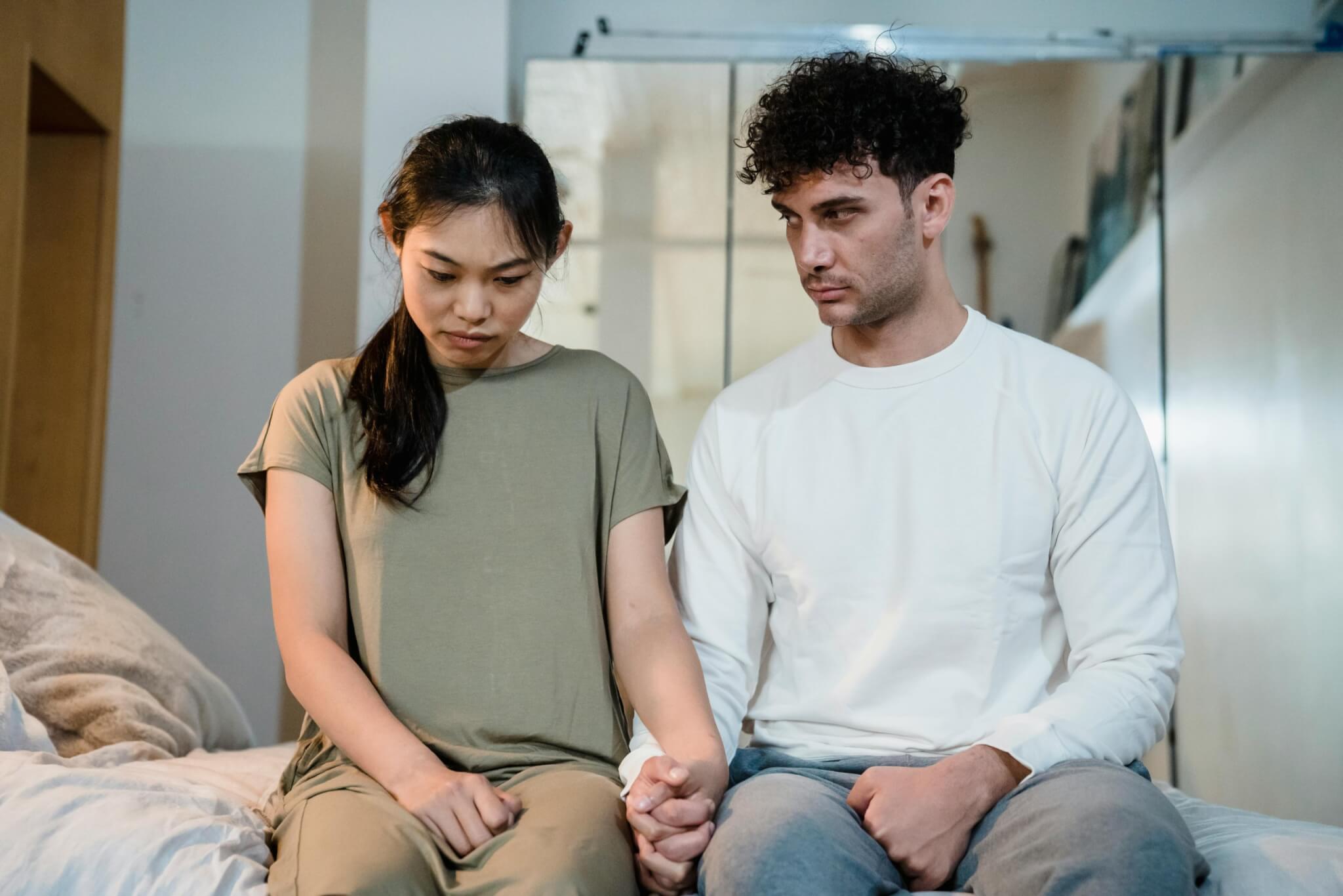 A couple sits on a bed, holding hands. The woman looks down with a somber expression, while the man gazes at her with concern. The room is softly lit, creating a calm atmosphere that belies their underlying relationship problems.