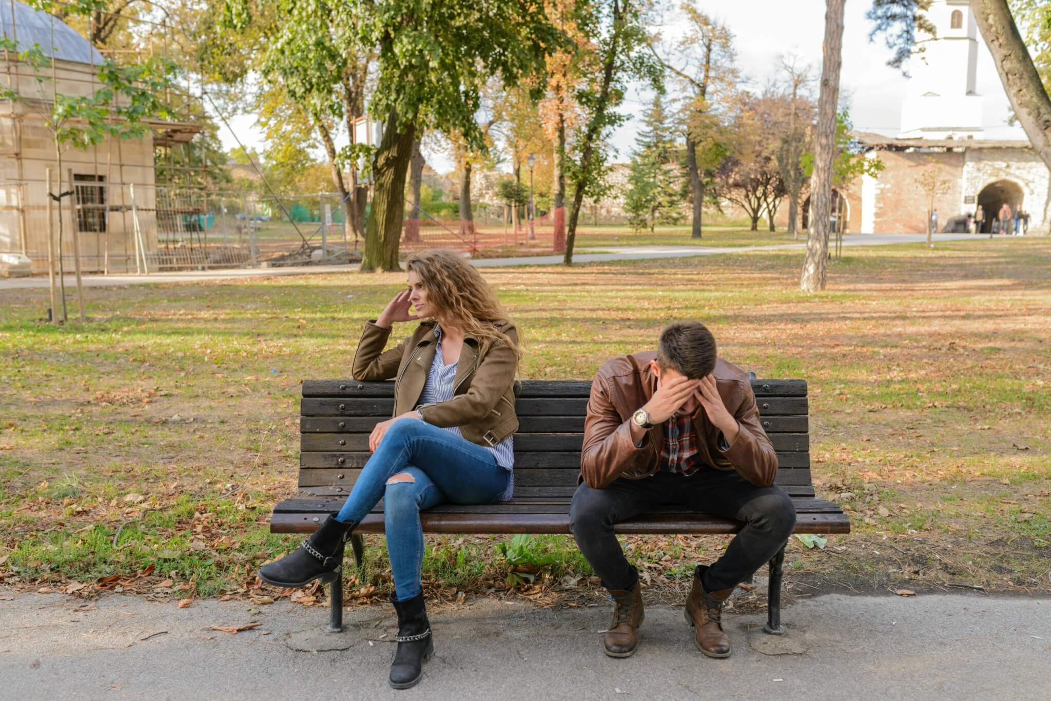 A woman and a man sit on a park bench, the weight of infertility casting a shadow. The woman looks away with a hand on her face, while the man is hunched over, his face buried in his hands. Trees and a building provide the muted backdrop to their shared silence.