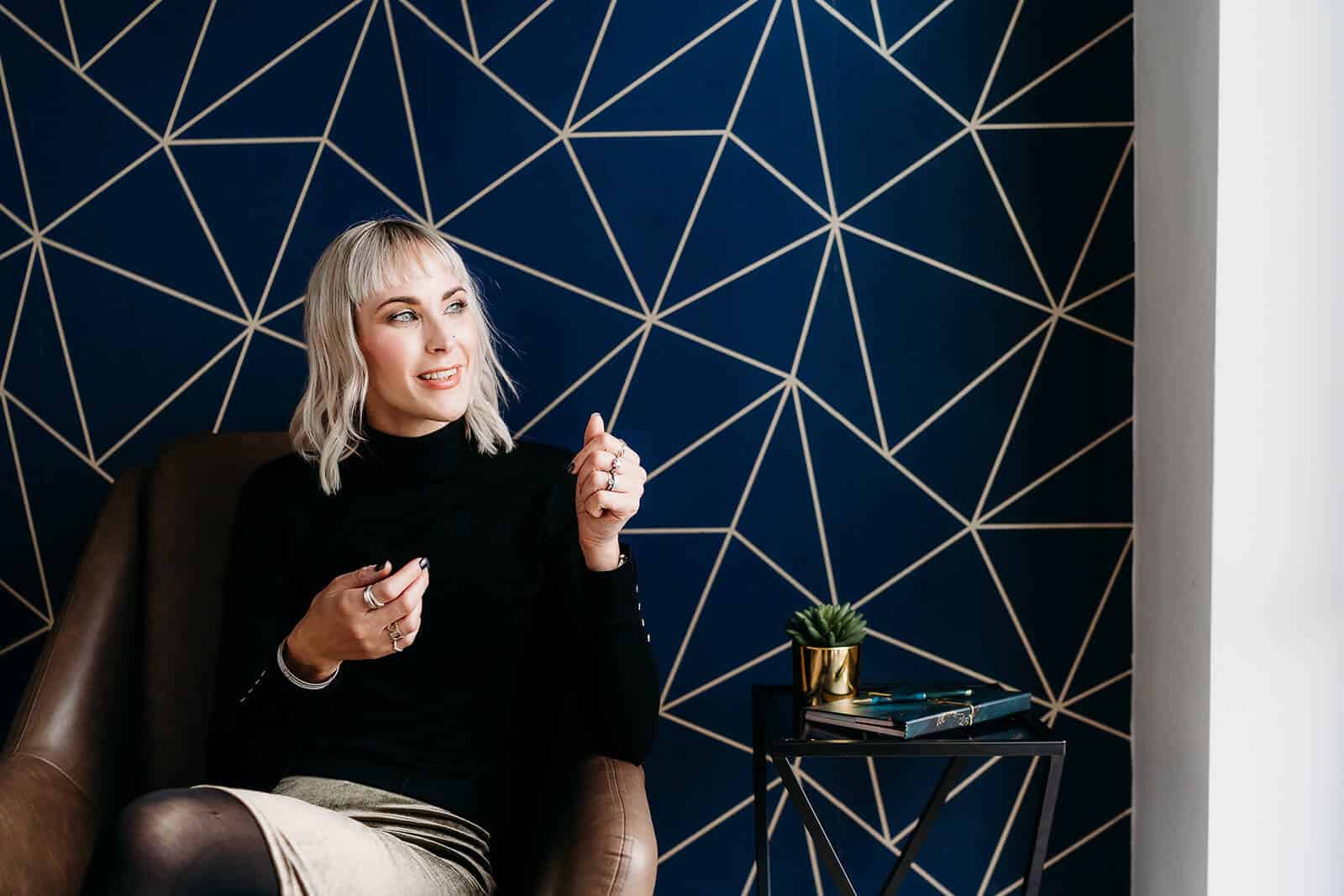 A person with short blonde hair sits in a chair against a geometric-patterned blue wall in Surrey. They are wearing a black top, holding a pen, and reflecting on therapeutic conditions. A small table with a plant and books is beside them.