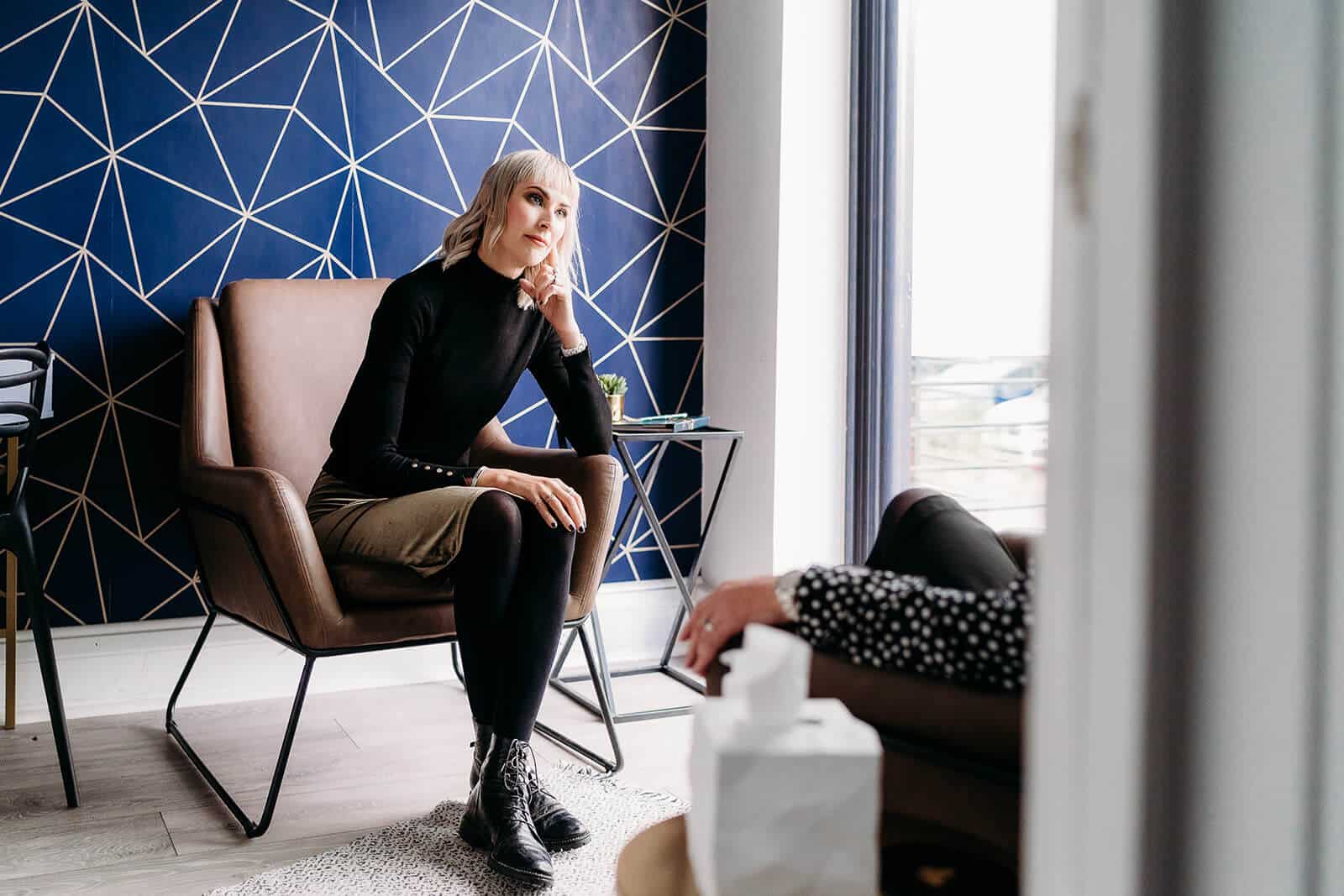A person with light hair sits thoughtfully in a brown chair, wearing a black top and brown skirt, contemplating conditions. Another individual is partially visible in the foreground. The backdrop features a blue accent wall with a geometric pattern, reminiscent of modern therapy spaces in Surrey.