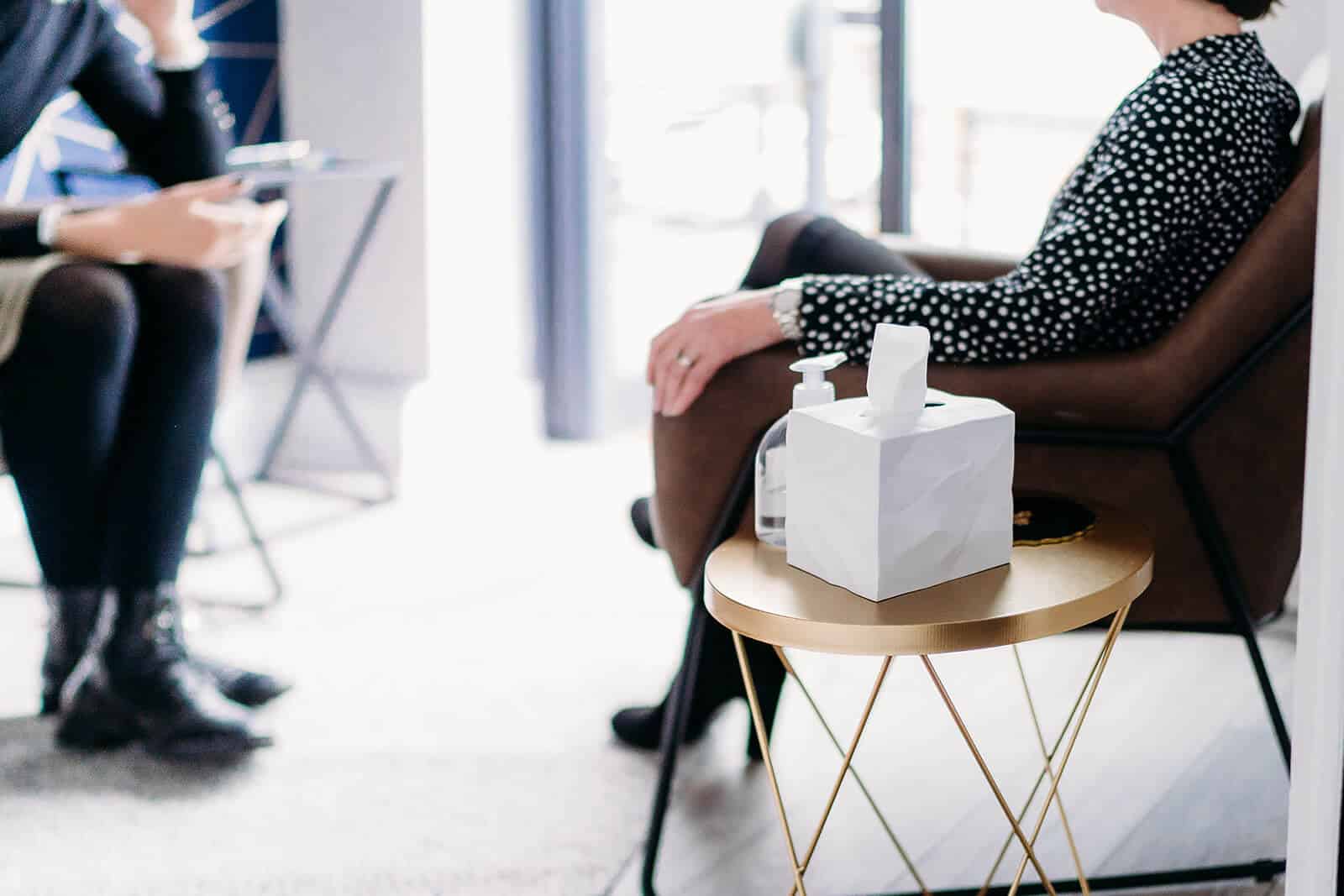 In a serene Surrey office setting, two people engage in comforting therapy. One wears a black and white polka dot outfit, seated on a brown chair. A gold table between them holds a tissue box and sanitizer bottle, ensuring hygiene is maintained amidst the session.