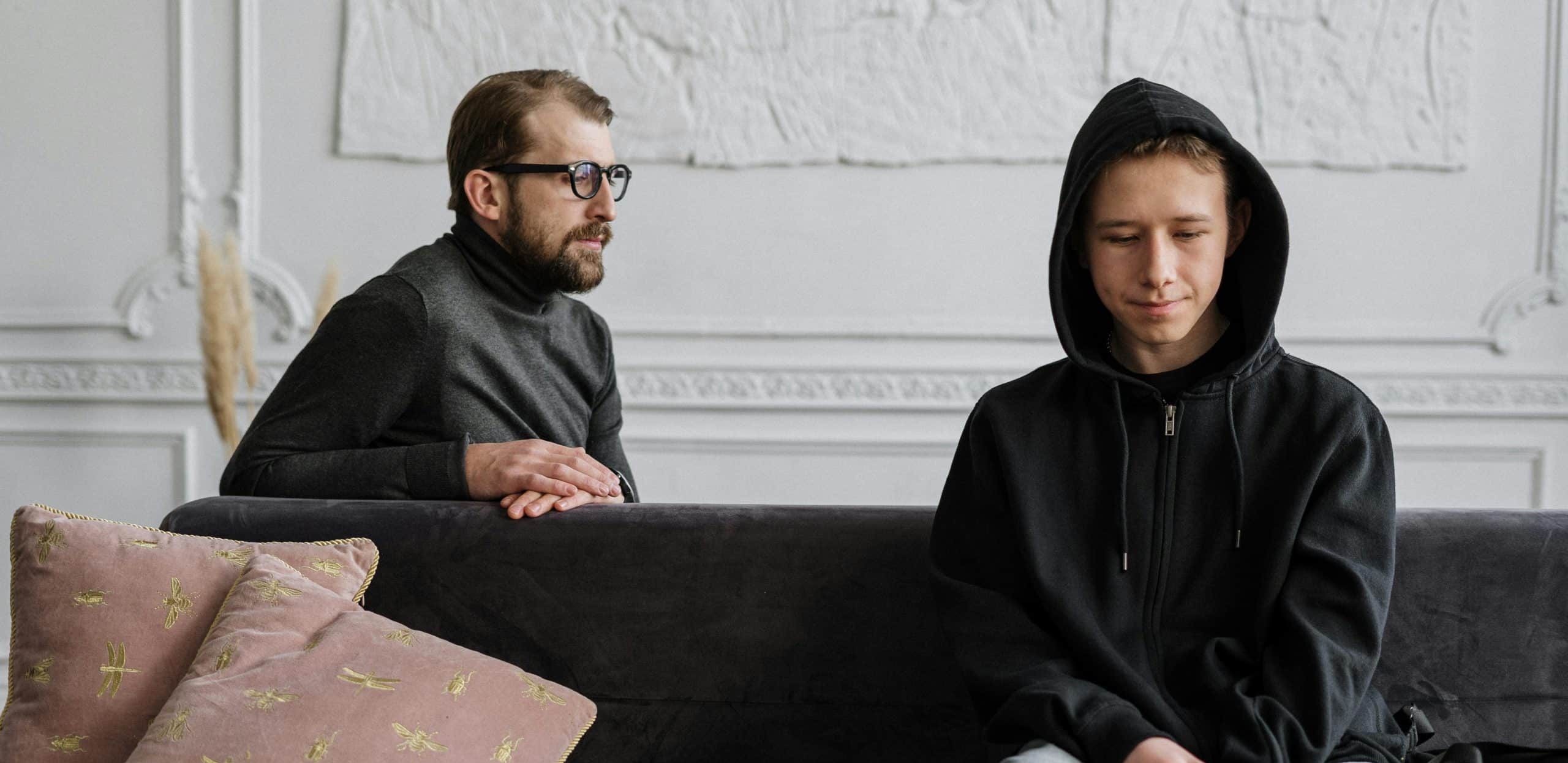 A man with glasses sits on a couch, contemplating the nuances of EMDR vs DBR while looking at a boy in a black hoodie sitting with a downcast expression. The room is bright with white walls and features decorative pillows on the couch.