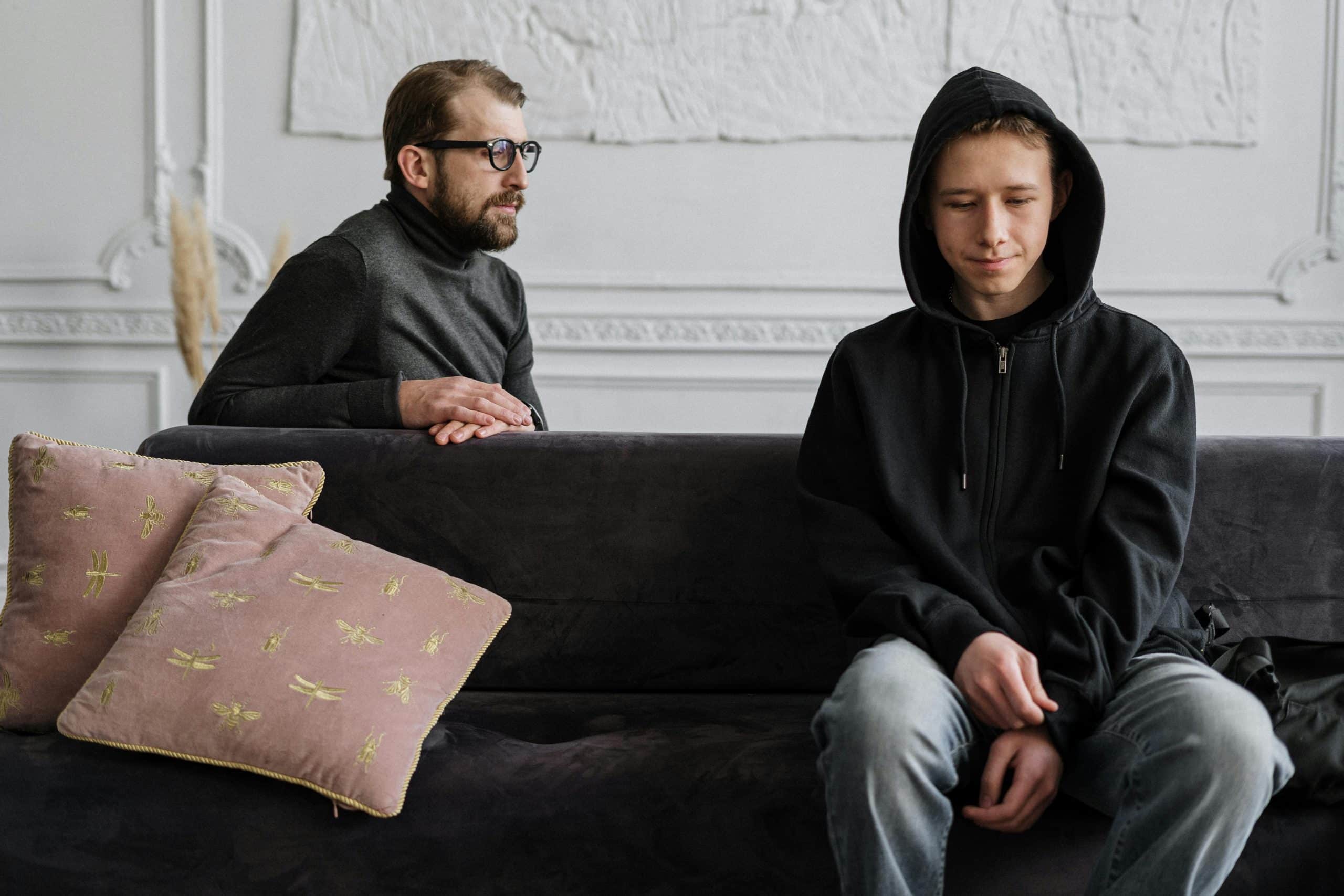 A man with glasses sits on a couch, contemplating the nuances of EMDR vs DBR while looking at a boy in a black hoodie sitting with a downcast expression. The room is bright with white walls and features decorative pillows on the couch.