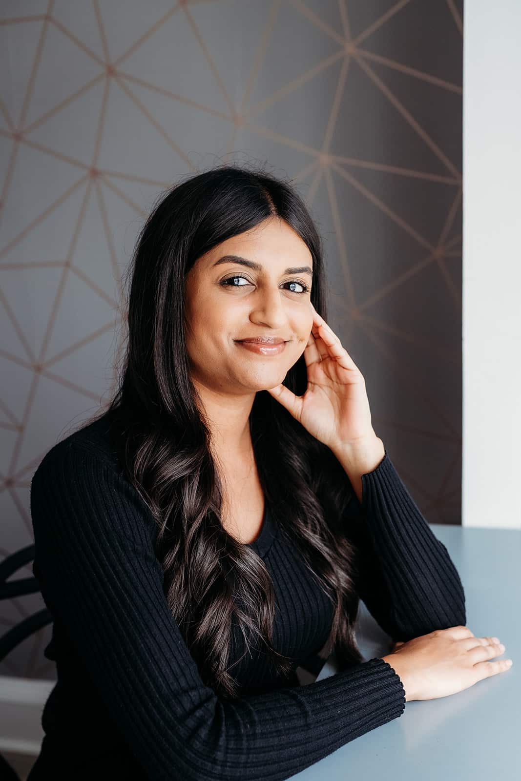 A woman with long dark hair and a black sweater sits at a table, resting her head on one hand and smiling softly. The background features a geometric pattern on a grey wall.
