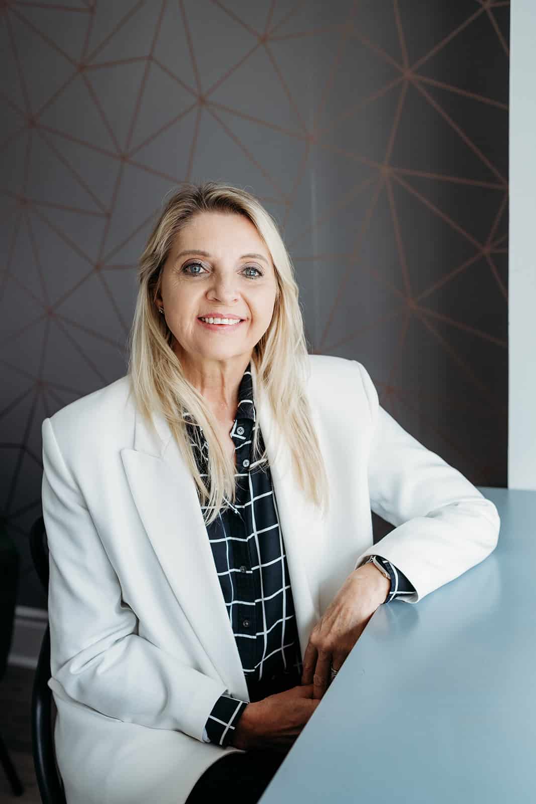 A woman with long blonde hair sits at a light blue table, smiling. She wears a white blazer over a black blouse with a white grid pattern. The background features a geometric design on gray walls.