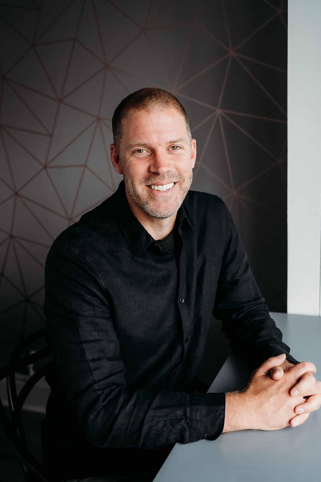Tom, a smiling man with short hair and a trimmed beard, sits at a gray table in front of a modern, geometric-patterned wall, wearing a black button-up shirt.