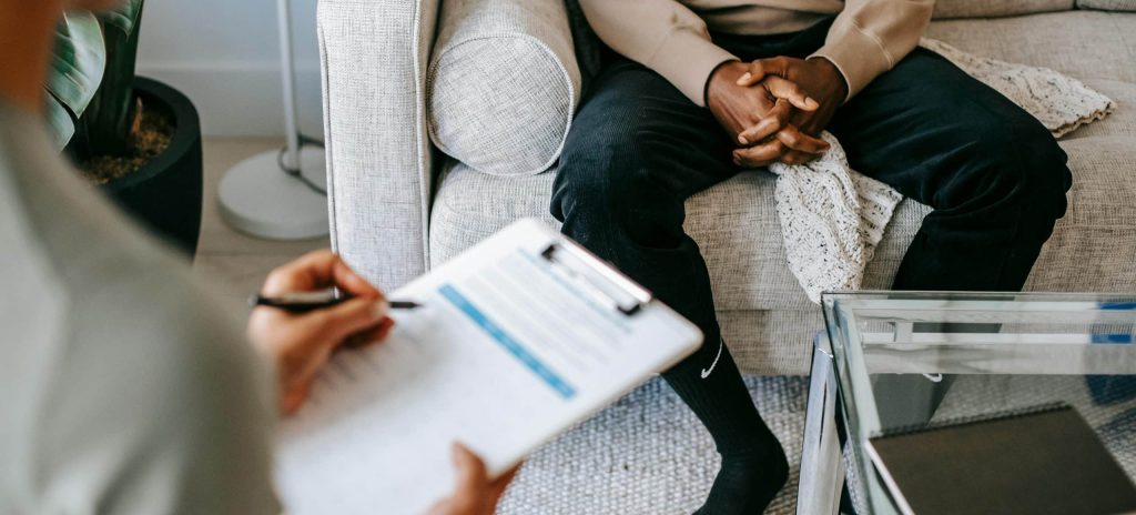 A person sits on a couch with hands clasped nervously while another, possibly a specialist in a particular therapy type, takes notes during the counseling session.