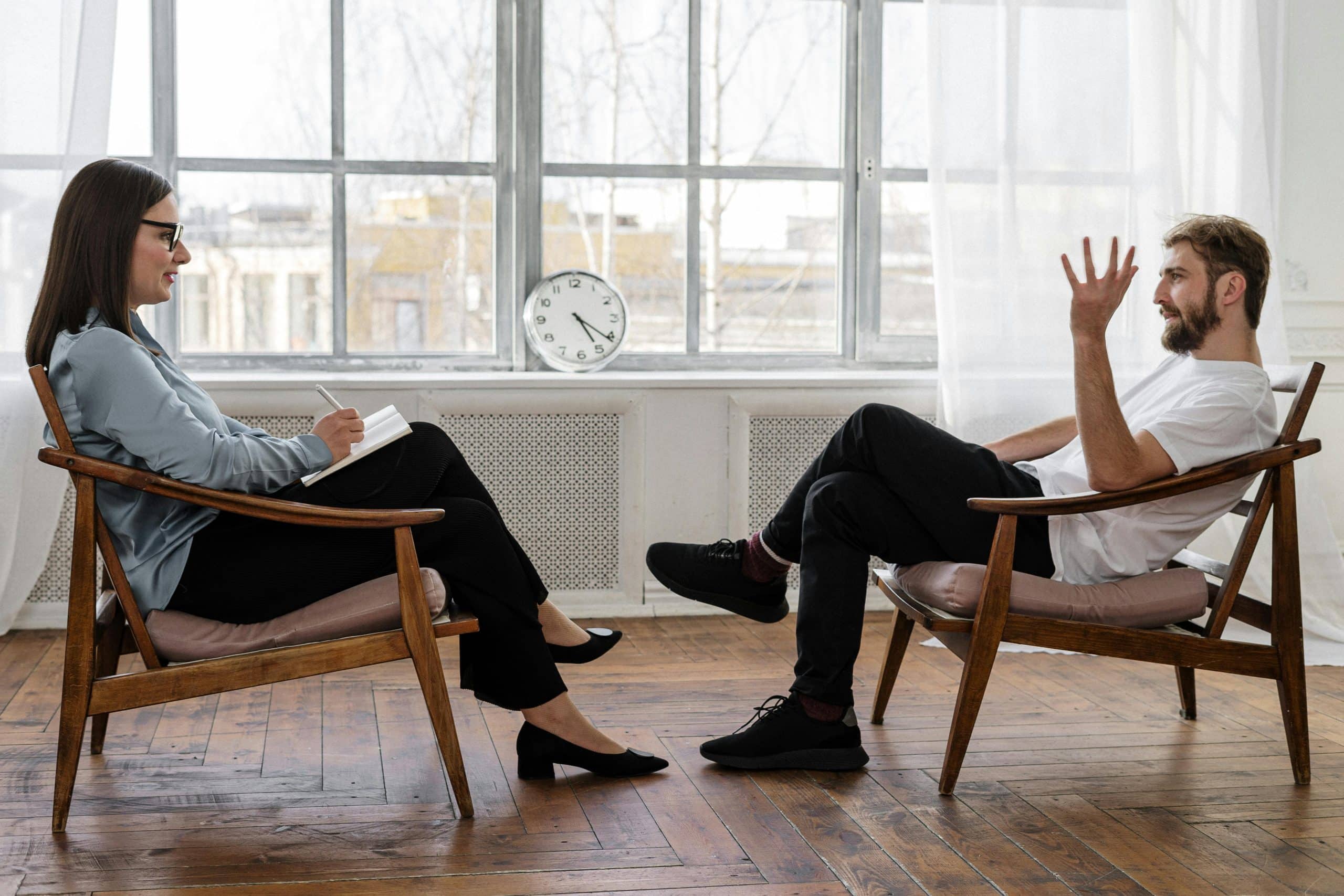 A woman with glasses sits taking notes while listening to a bearded man gesturing during a conversation about Therapy Type in a bright, modern room with large windows and a clock on the radiator behind them.