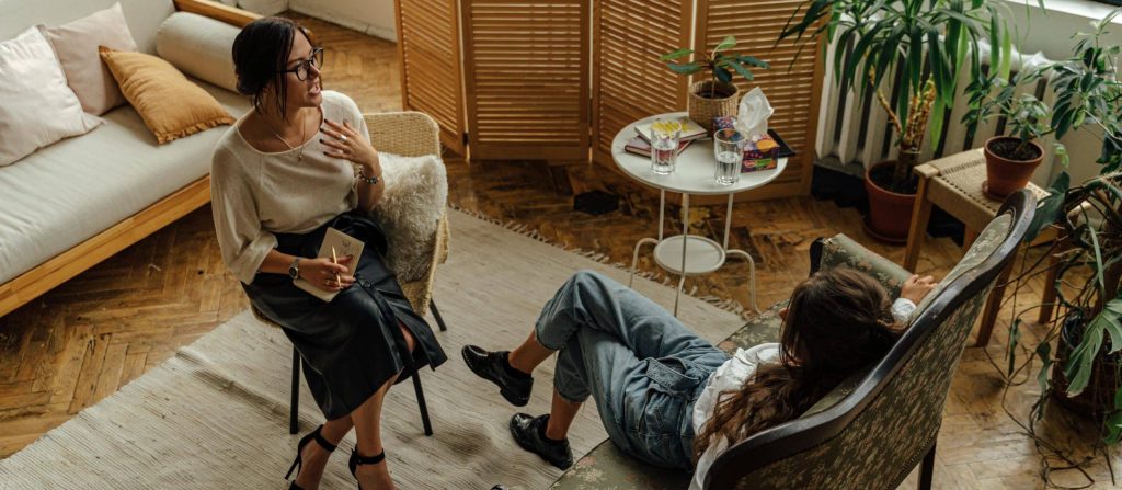 Two women sit in a cozy living room with plants, one on a sofa holding a notebook, the other reclining on a patterned couch. A small round table with books and drinks sits between them.
