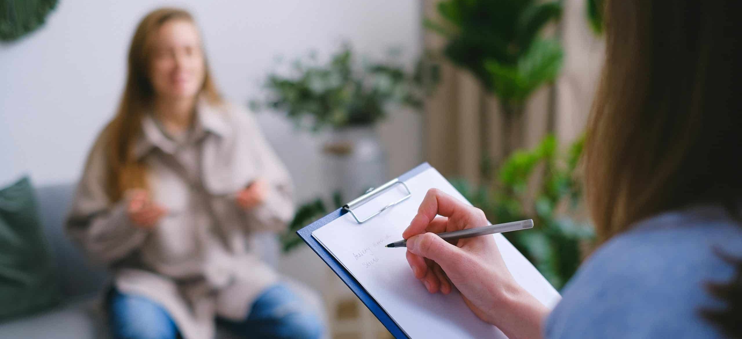 A person holding a clipboard and taking notes sits across from another person, blurred in the background, during a Therapy Type session in a room with plants and soft lighting.