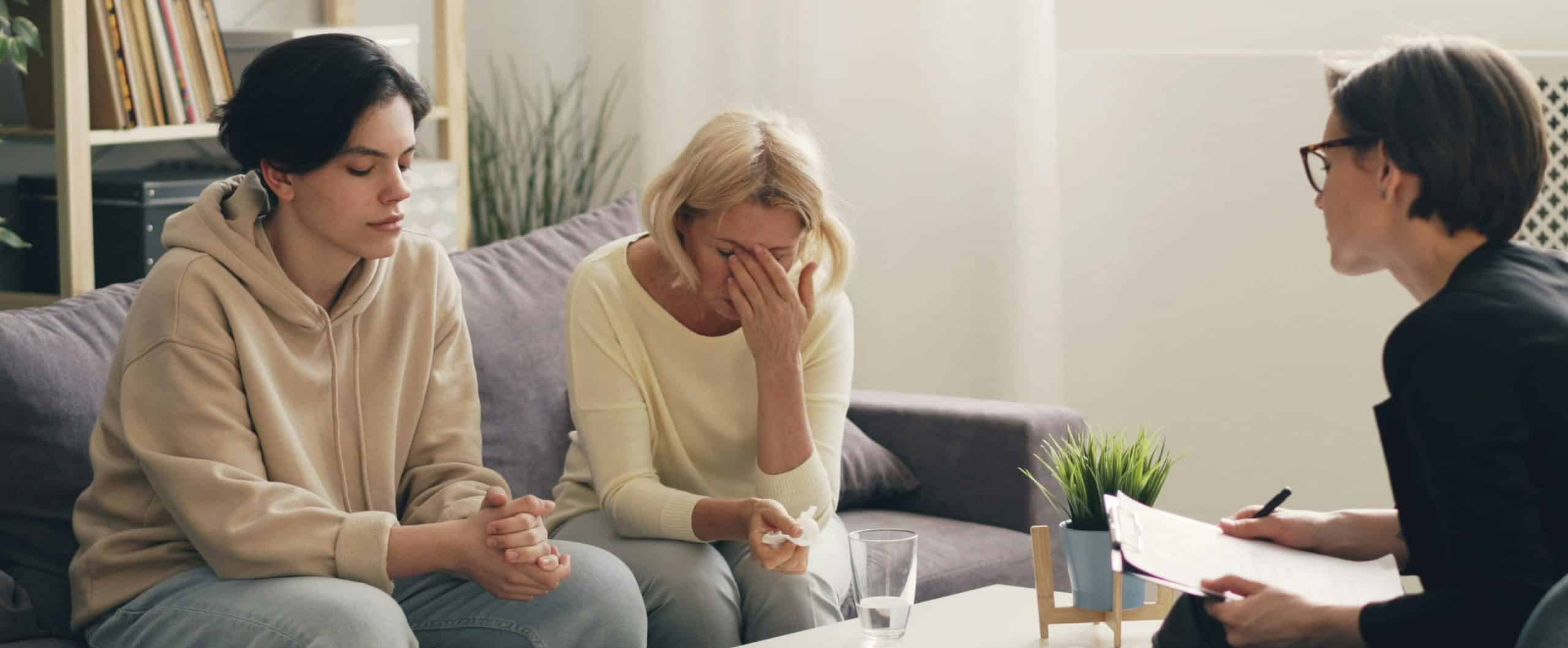 Two women sit on a couch, one with eyes closed and the other covering her face, looking distressed. Across from them, a person holding a clipboard suggests this is a therapy session focused on a specific Therapy Type.