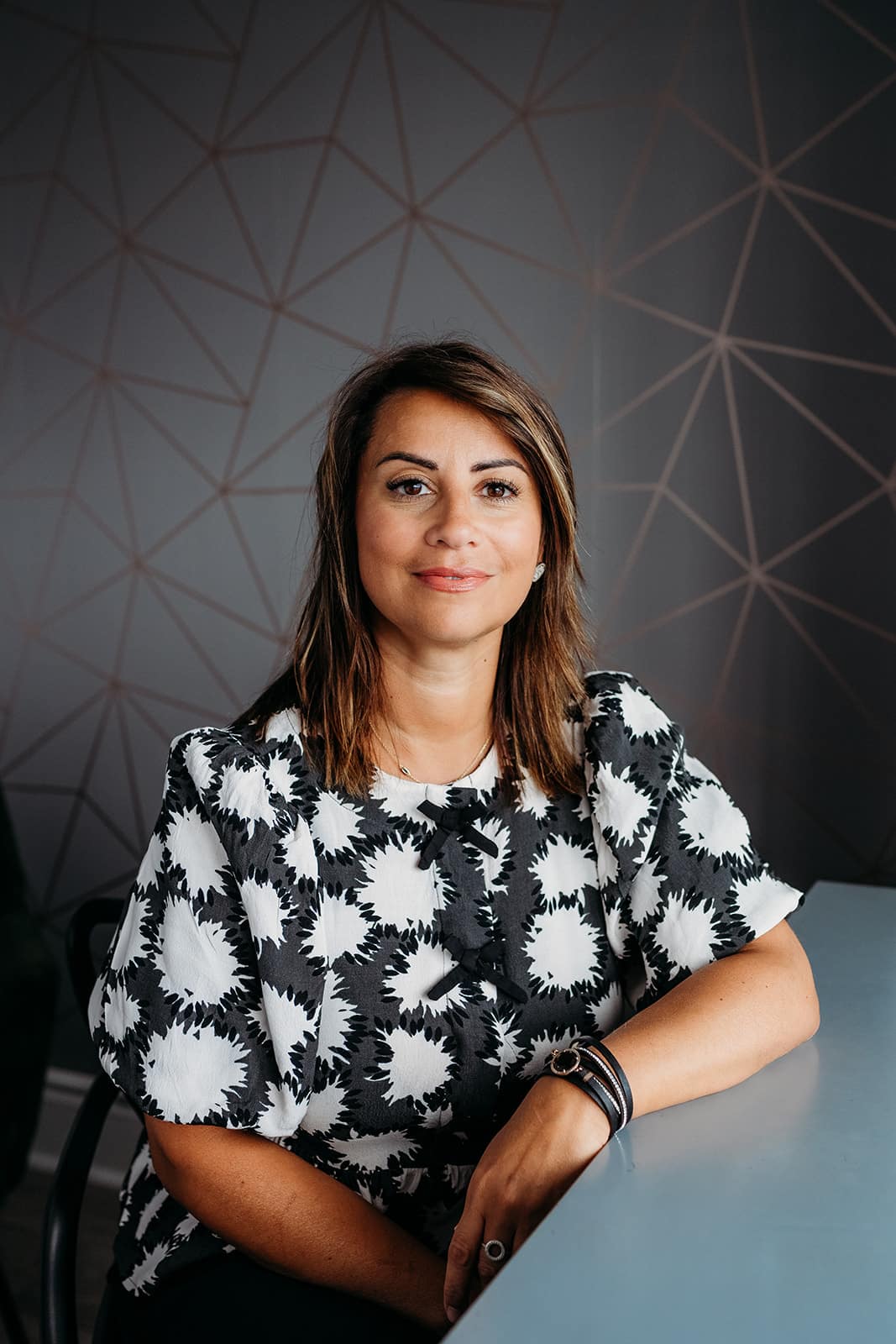 A woman with shoulder-length brown hair, wearing a black and white patterned blouse, sits at a table with her arms resting on it, smiling softly in front of a geometric-patterned wall.