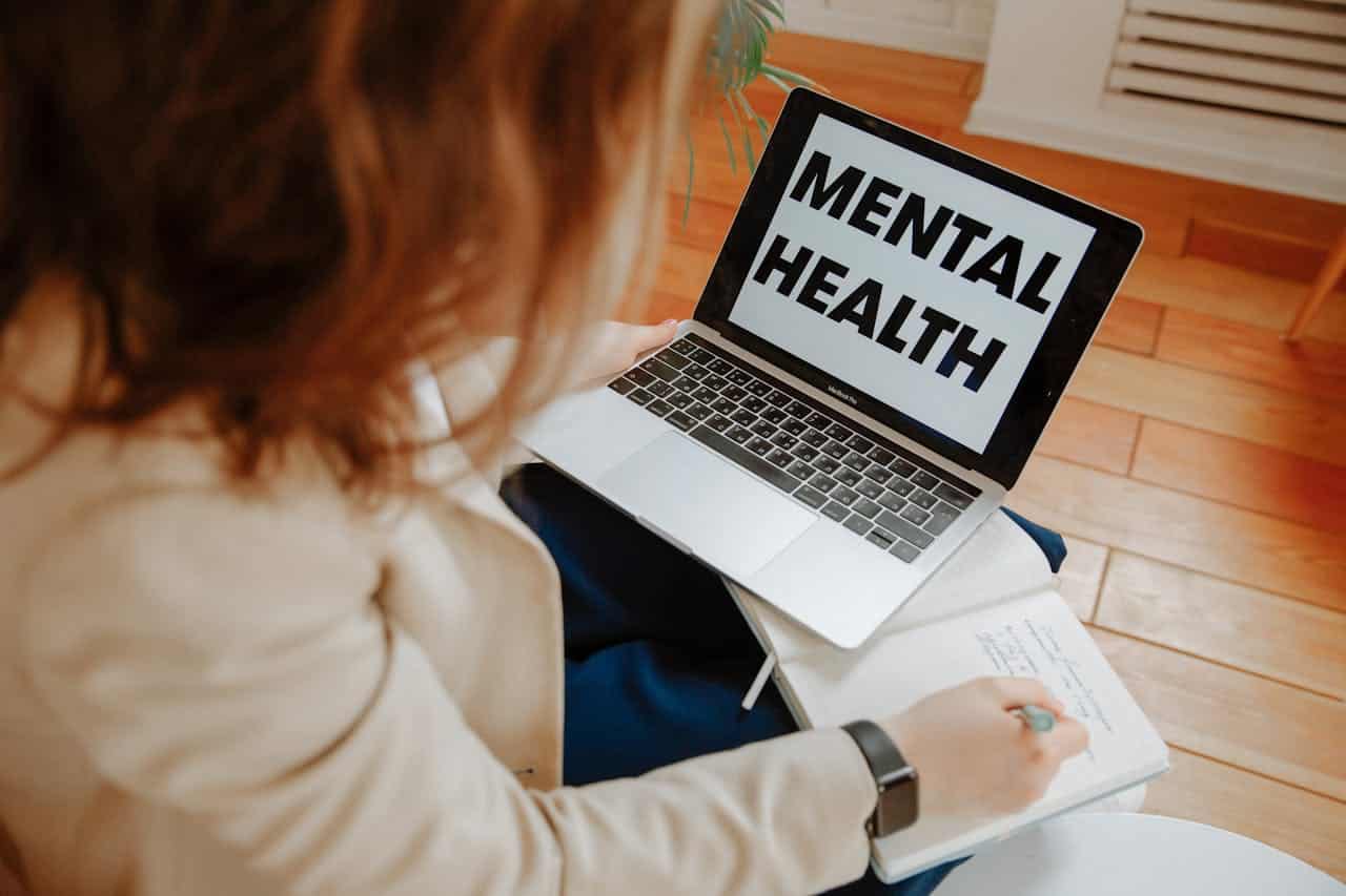 A person holding a notebook and pen sits at a desk, looking at a laptop screen displaying the words MENTAL HEALTH in bold black letters—a scene reflecting workplace stress & burnout support.