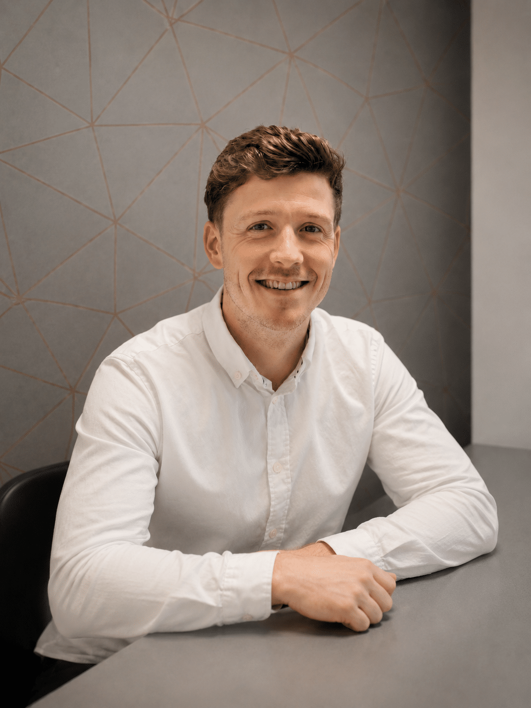 A man with short brown hair, wearing a white button-up shirt, sits at a grey desk and smiles at the camera. The background features a grey wall with a geometric line pattern.