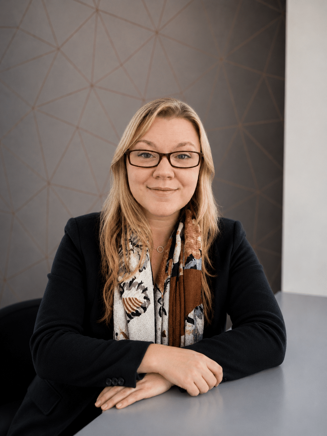 A woman with long blonde hair and glasses sits at a table, smiling. She wears a dark blazer and a patterned scarf. The background features a grey wall with a geometric pattern.