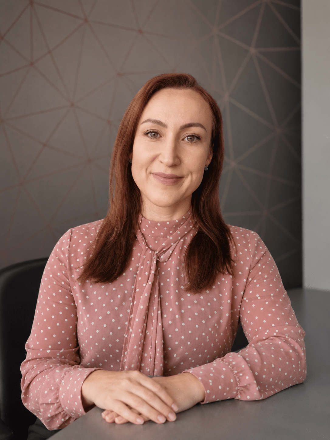 A woman with straight, reddish-brown hair sits at a table, smiling gently. She wears a pink blouse with white polka dots and rests her hands on the table. The background features a geometric patterned wall.