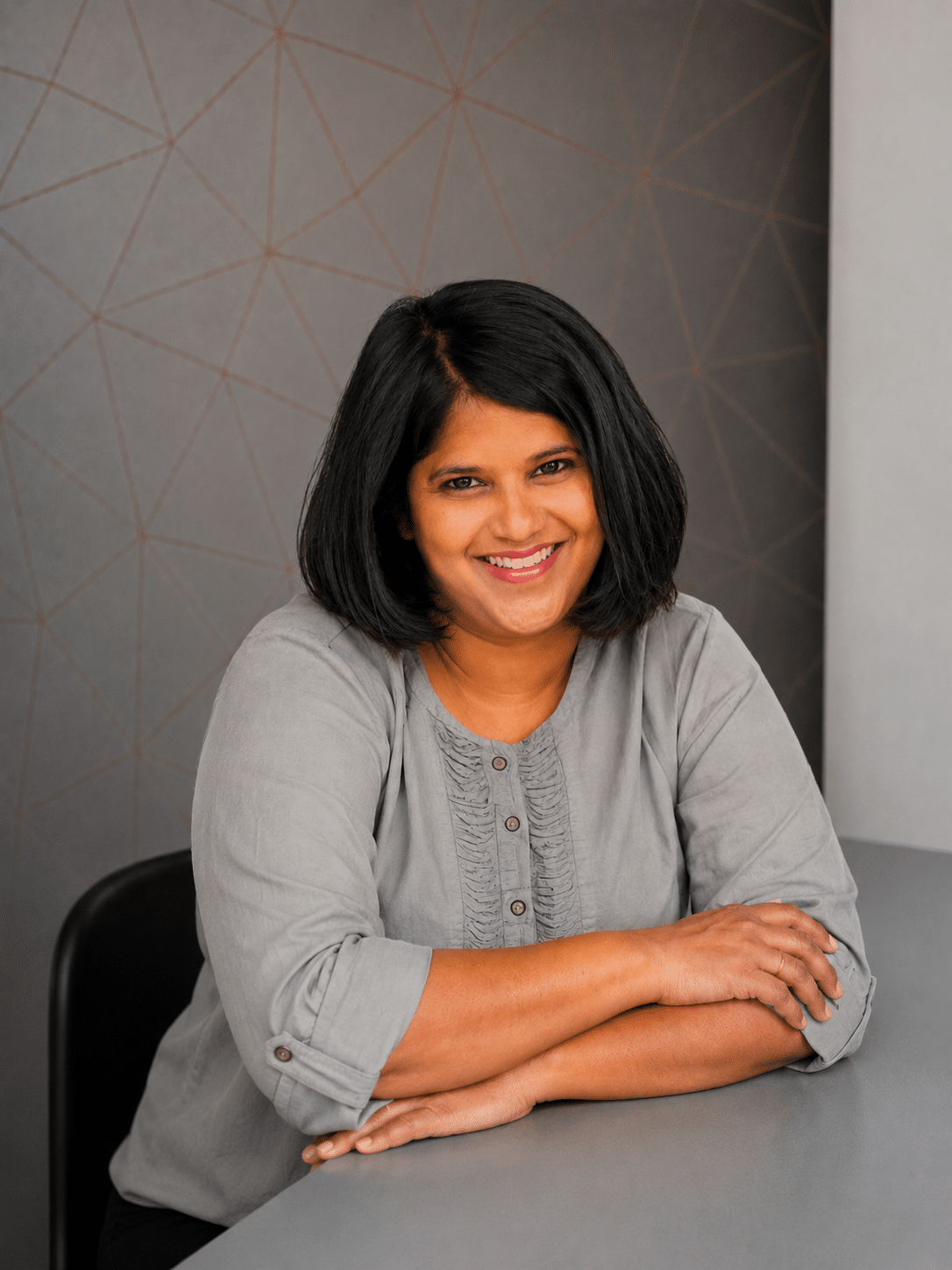 A woman with shoulder-length black hair, wearing a light grey blouse, sits at a table with her arms crossed and smiles at the camera. The background features a modern geometric pattern in grey tones.