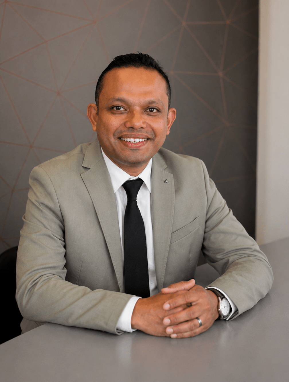 A man in a light grey suit and black tie sits at a desk, smiling at the camera. The background features a grey wall with a geometric line pattern.