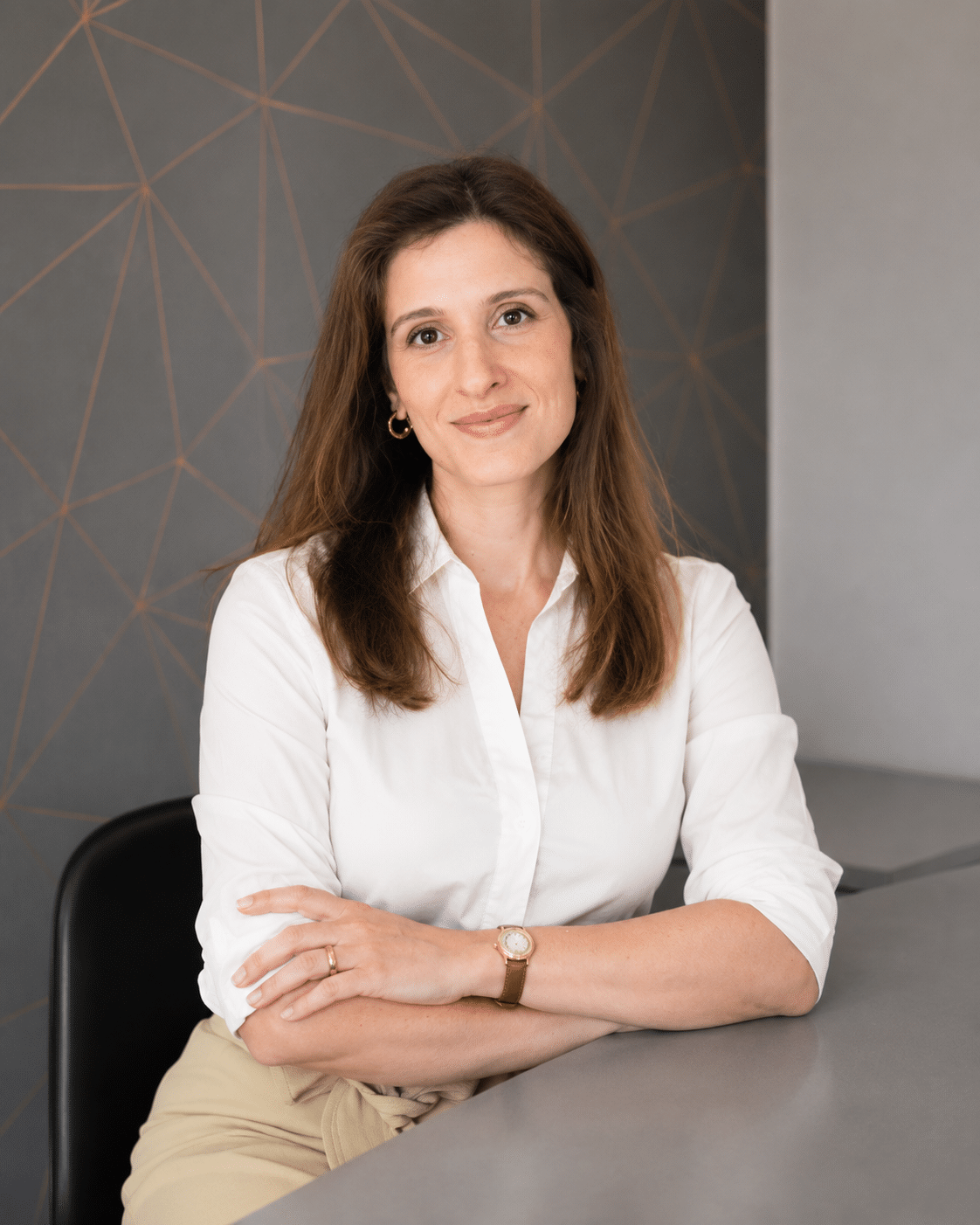 A woman with long brown hair, wearing a white blouse and beige trousers, sits at a table with her arms crossed, smiling slightly. The background features a grey wall with a geometric gold line pattern.